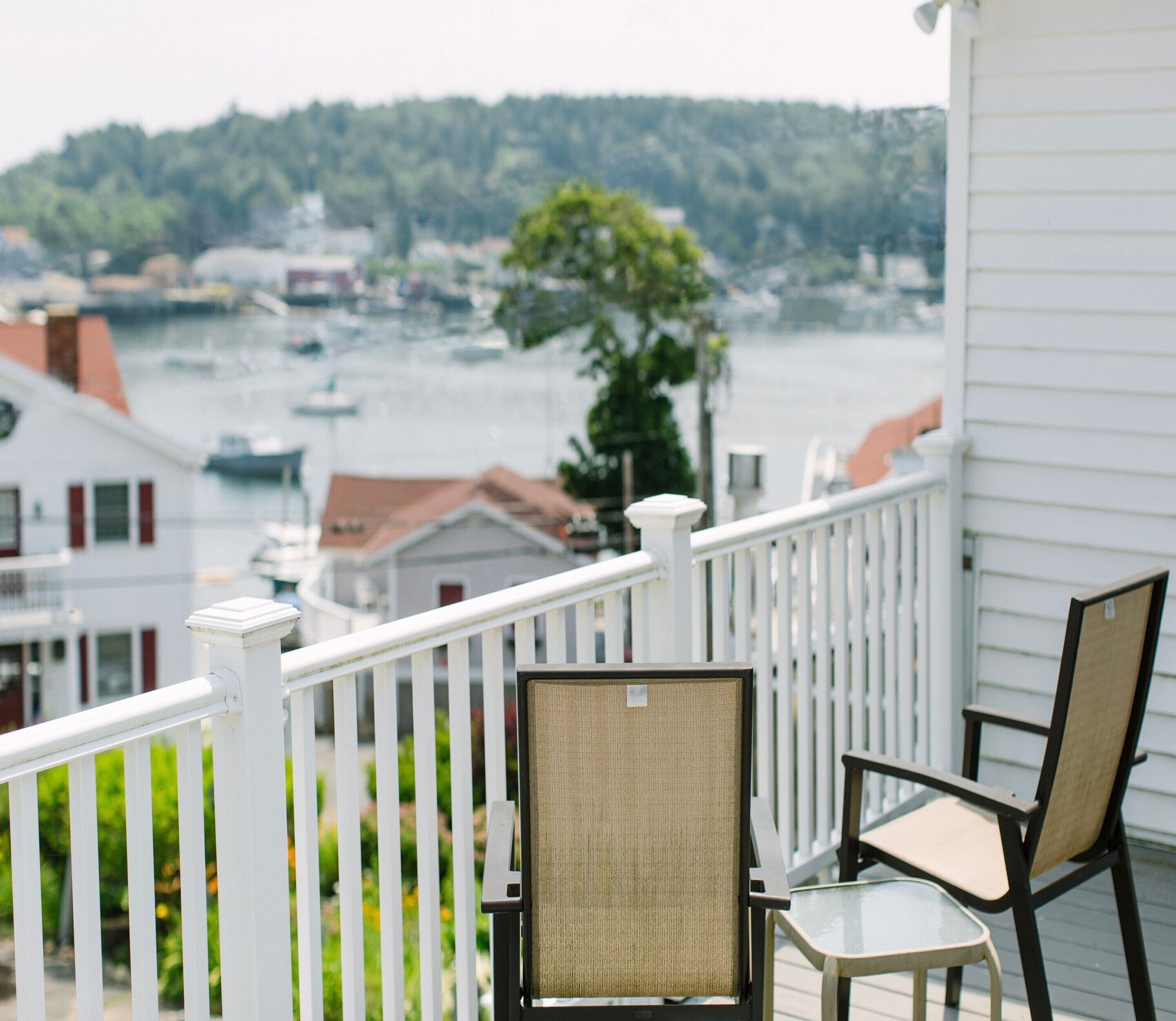 A balcony with white railings, two chairs, and a view of a lake with boats and buildings in the background.