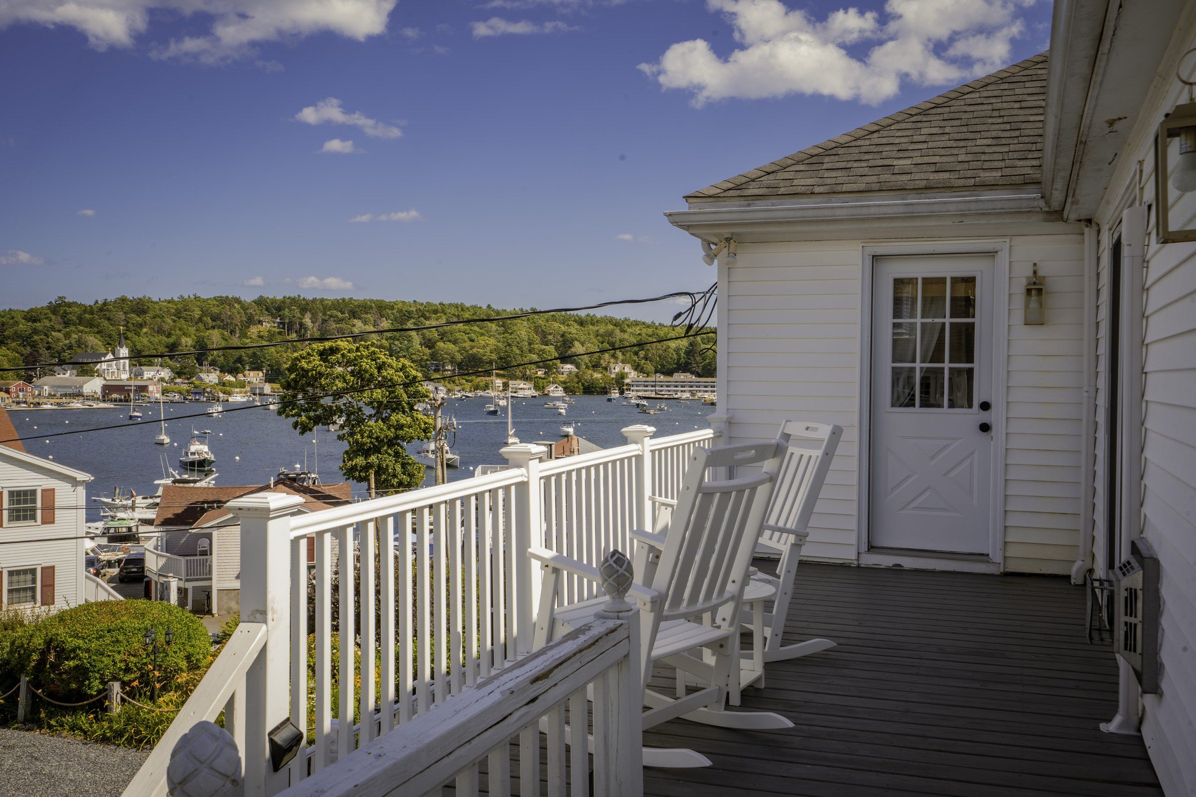 A white building with a wooden deck and rocking chairs, overlooking a marina with boats.
