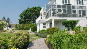 A white two-story house with a balcony, surrounded by a garden with various plants and flowers. - Admirals Quarters Inn (Boothbay Harbor)