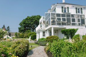 A white two-story house with a balcony, surrounded by a garden with various plants and flowers.