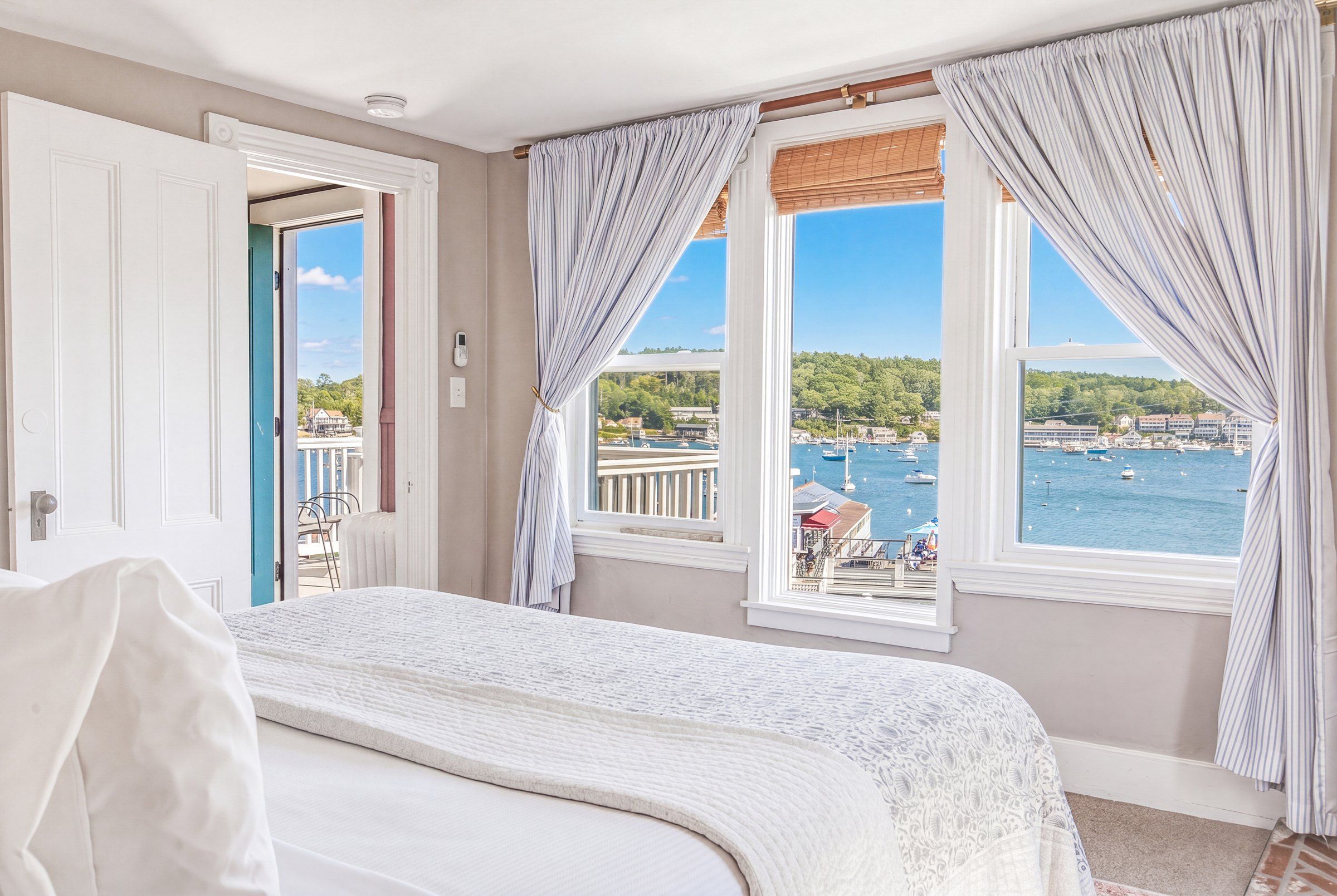 Quarto de hotel com cama, duas janelas com cortinas, vista para o mar e varanda com cadeiras.