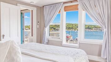 Quarto de hotel com cama, duas janelas com cortinas, vista para o mar e varanda com cadeiras.