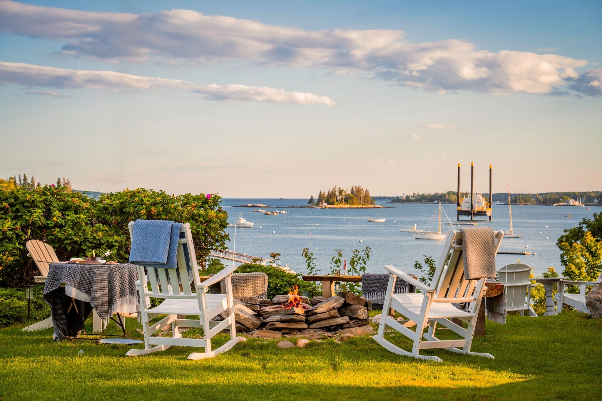 A lakeside view with two rocking chairs facing a fire pit, surrounded by greenery and overlooking a calm body of water with boats.