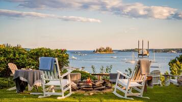 A lakeside view with two rocking chairs facing a fire pit, surrounded by greenery and overlooking a calm body of water with boats.