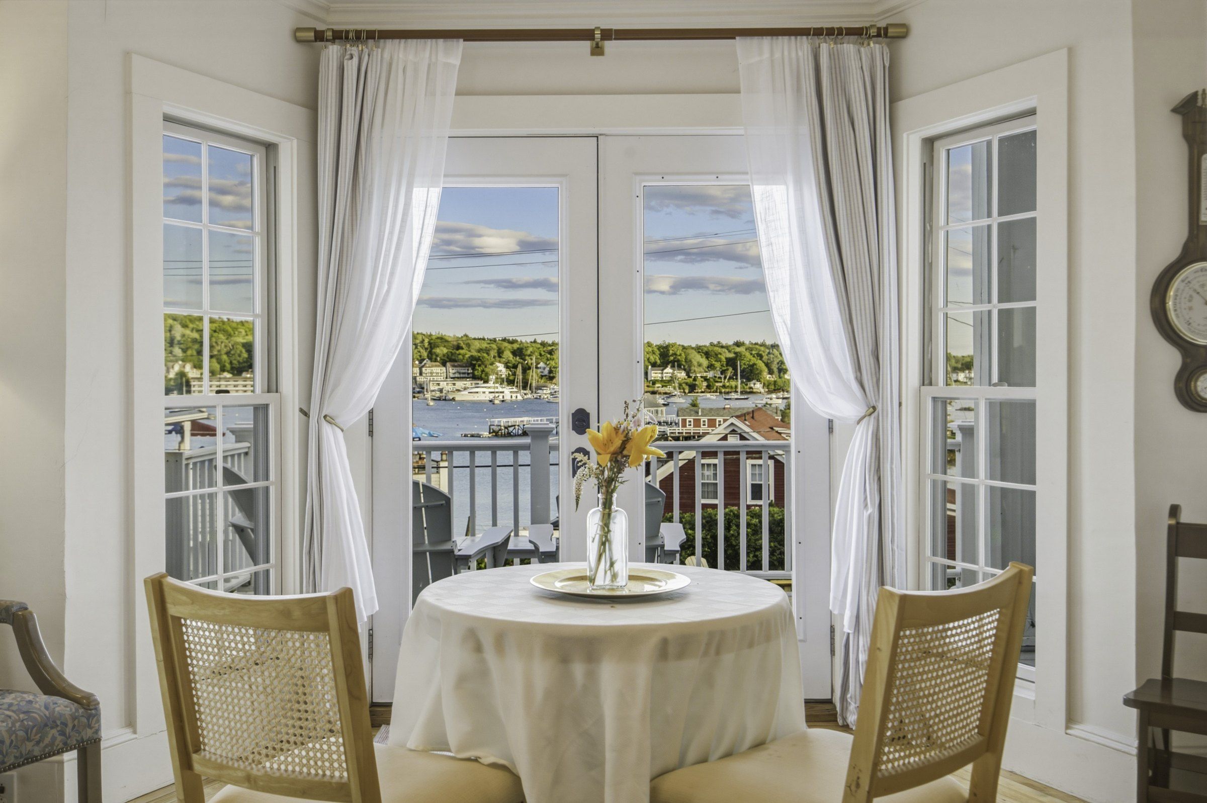 A room with a round table, two chairs, a vase with flowers, and a view of a marina through large windows with white curtains.