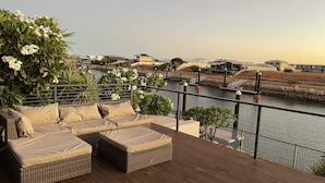 A wooden deck with wicker furniture and a view of a river and buildings in the background. - Gecko - On the Marina With Pool & Private Jetty, 4 Kestrel Place, Exmouth (Exmouth)