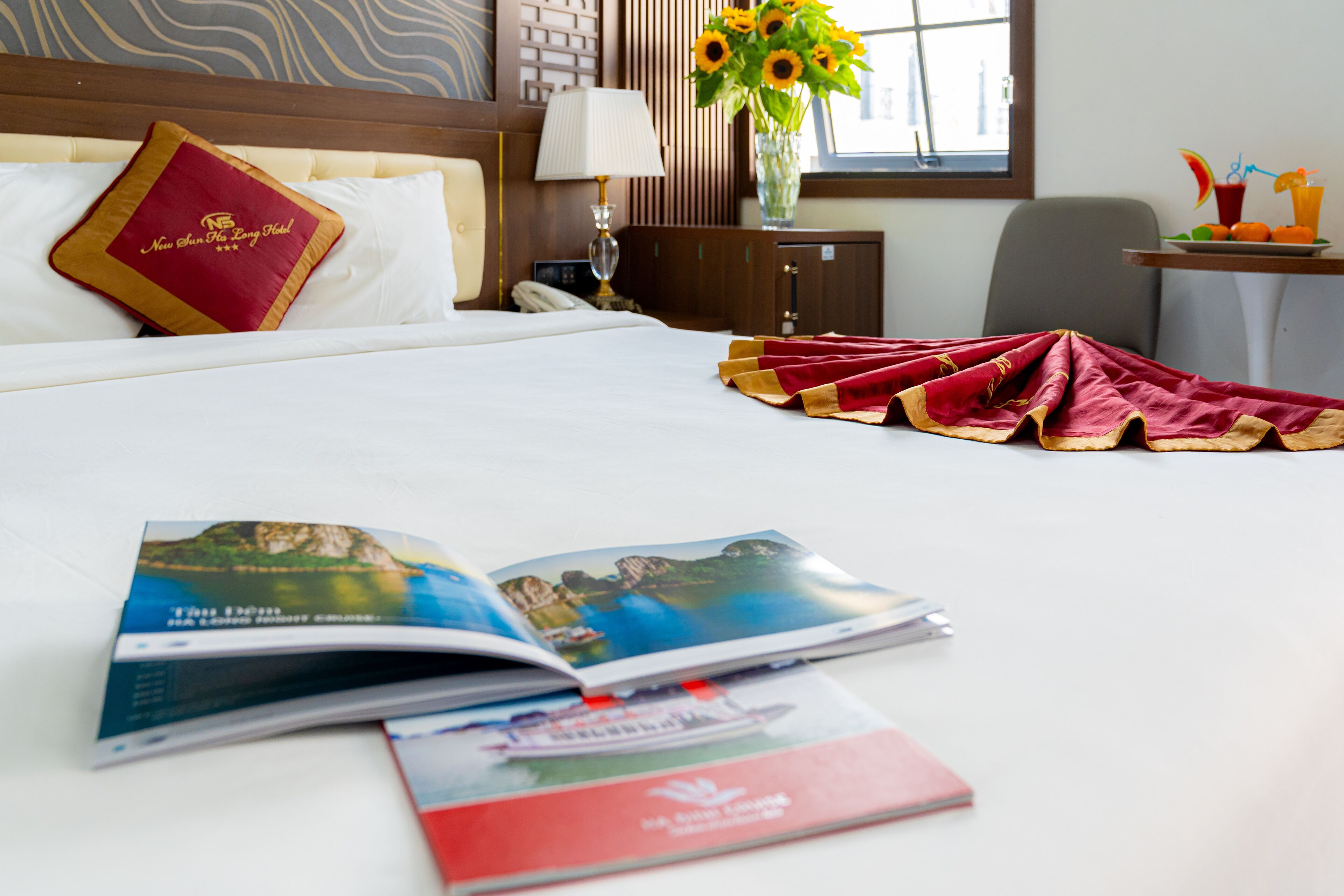 A hotel room with a neatly made bed, a bedside table with a lamp, a vase with sunflowers, and a magazine on the bed.