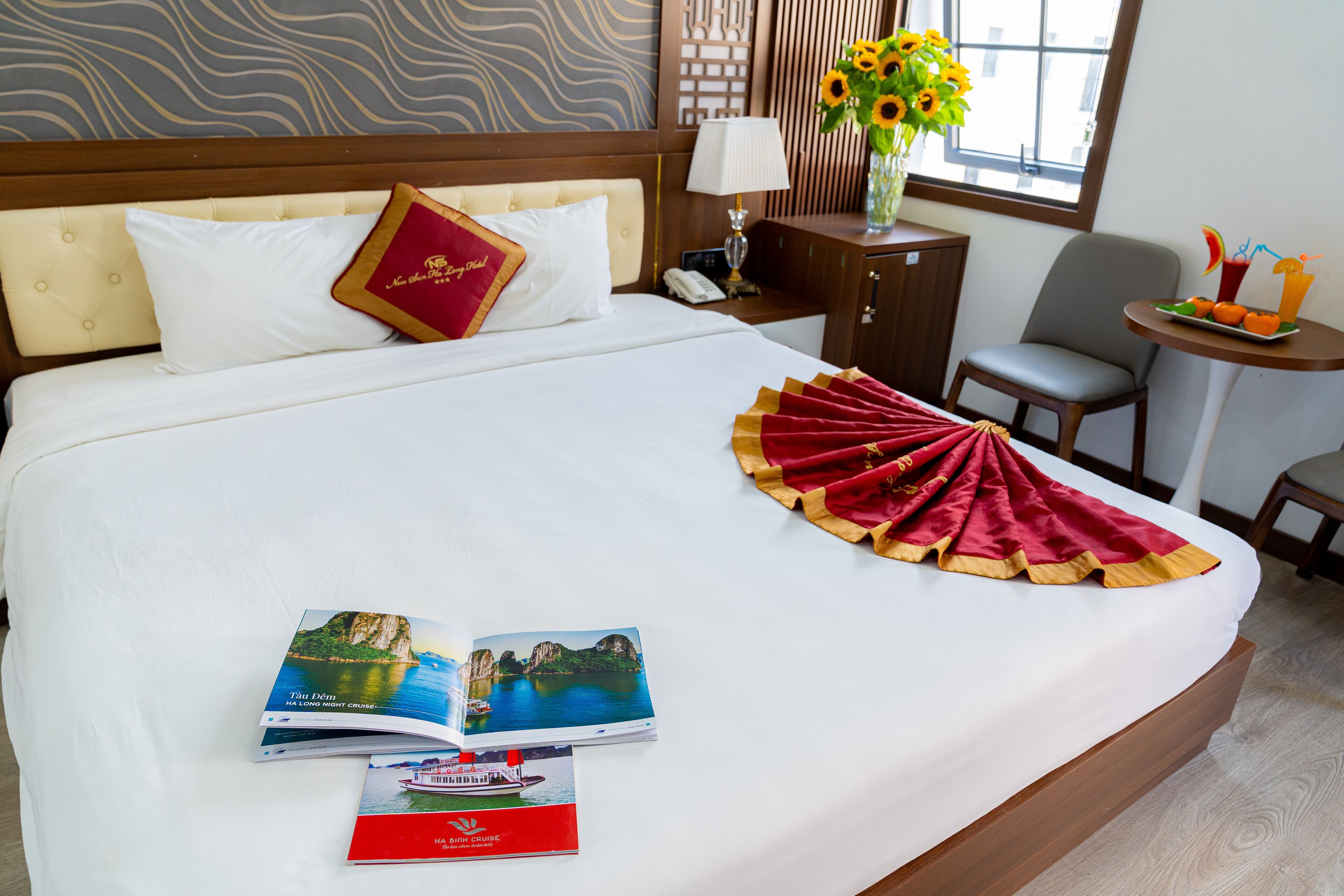 A hotel room with a large bed, a bedside table with a lamp, a chair, a small table with a fruit tray, and a window with a view of the city.