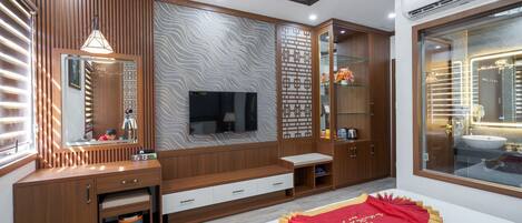 Una habitación de hotel moderna con un tocador de madera, un televisor de pantalla plana, un espejo grande y un baño con mampara de vidrio.