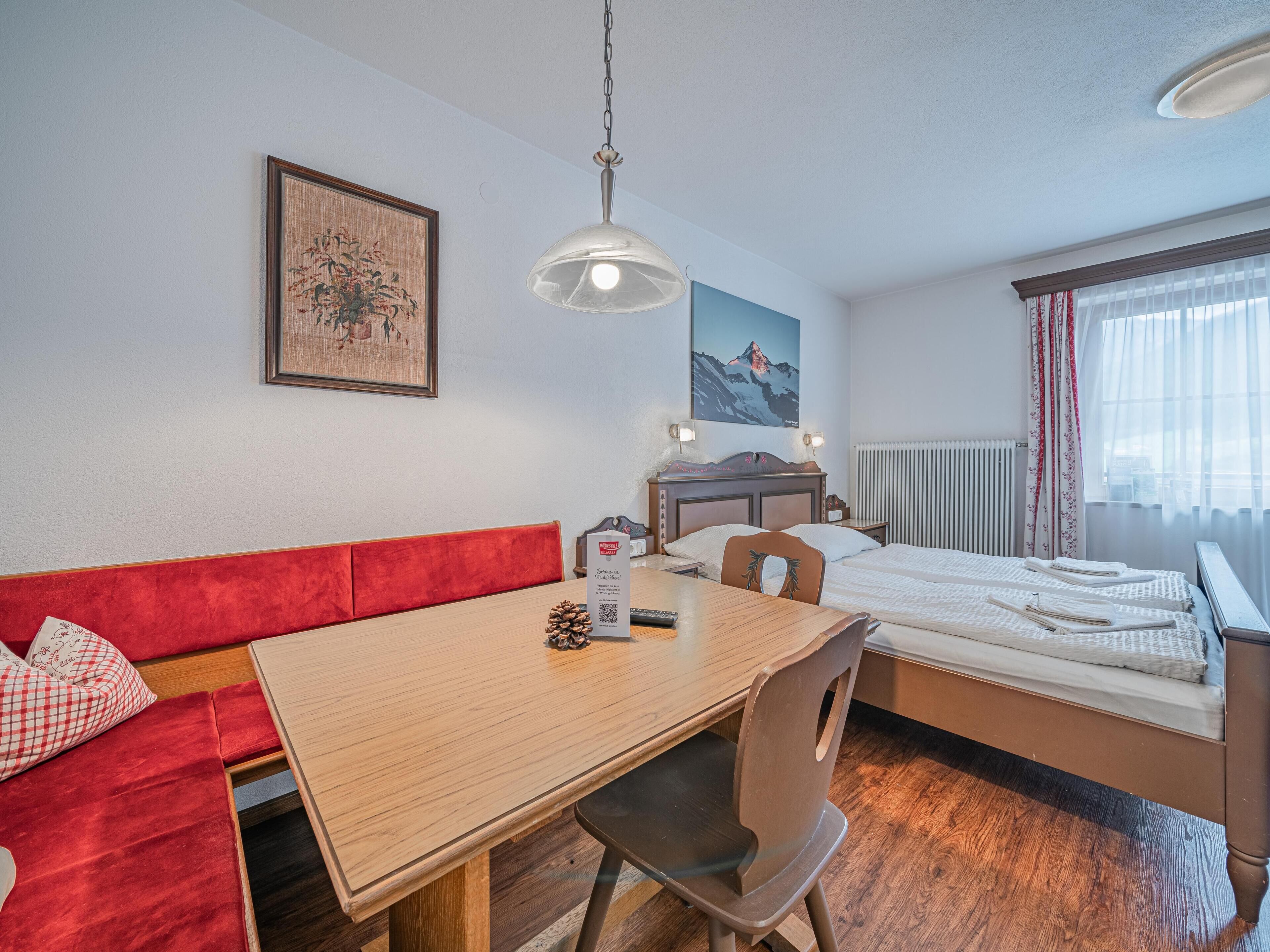 Une chambre d’hôtel avec une table à manger en bois, un banc recouvert d’un tissu rouge, un lit simple, une petite table de chevet, un tableau accroché au mur et une fenêtre avec des rideaux légers.