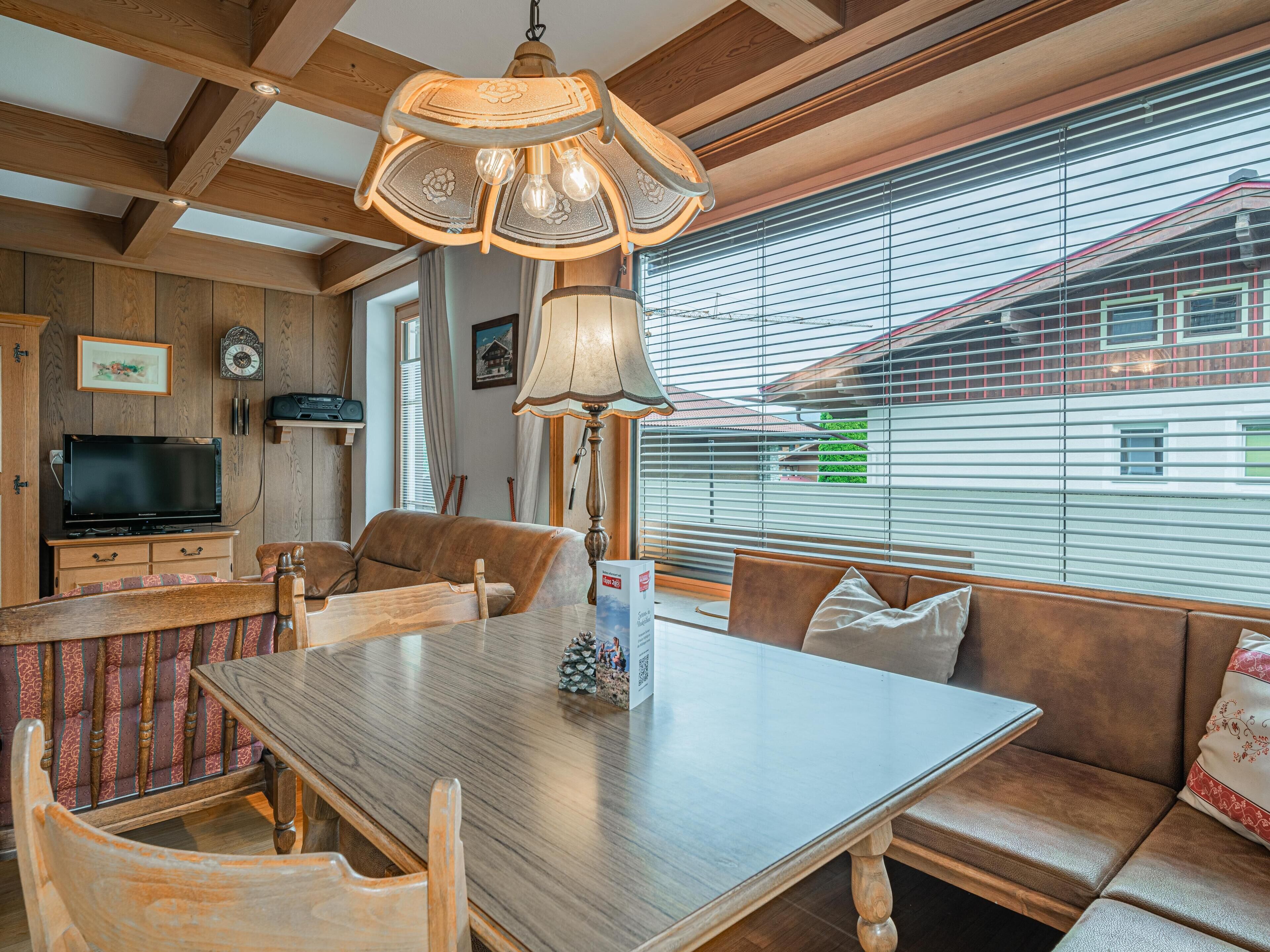A cozy living room with a wooden dining table, cushioned seating, a television, and a large window with blinds.