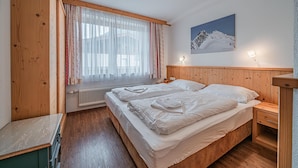 A bedroom with a wooden headboard, a bed with white linens, a radiator, a window with sheer curtains, and a wall-mounted light.