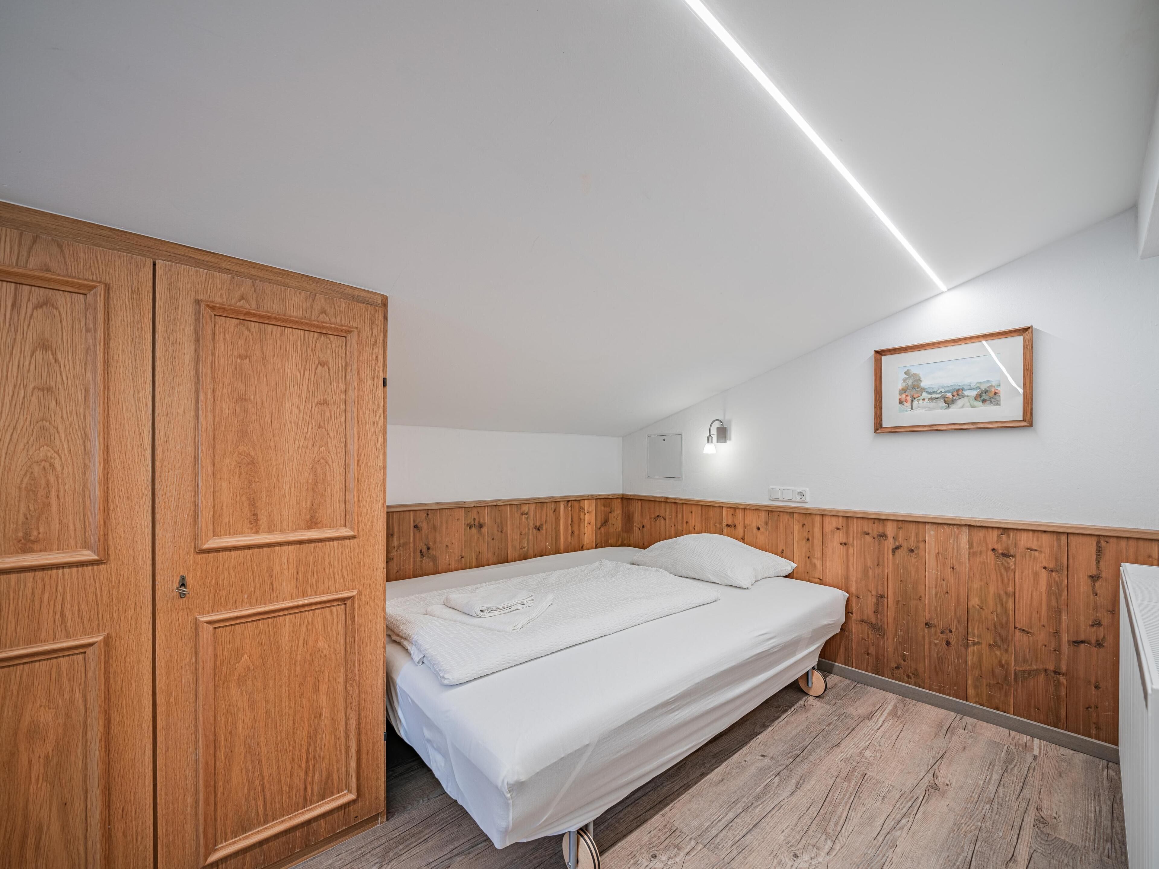 Ein Schlafzimmer mit einem Holzschrank, einem Einzelbett mit weißer Bettwäsche, einer Wandleuchte und einem gerahmten Bild an der Wand.