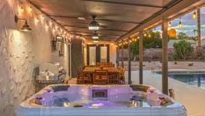 A hot tub with a wooden table and chairs, surrounded by string lights and a pool.