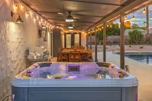 A hot tub with a wooden table and chairs, surrounded by string lights and a pool.