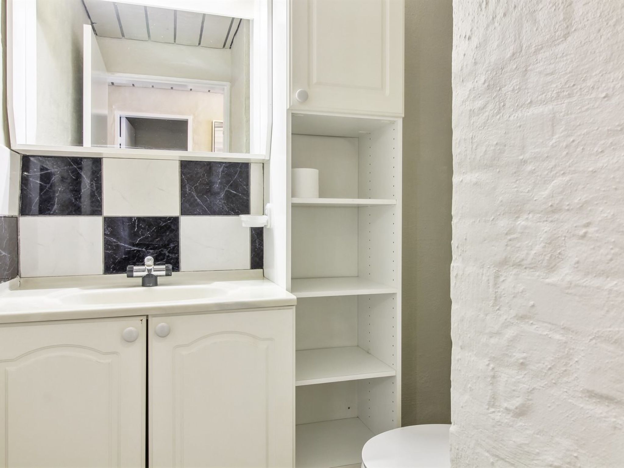 Un baño con tocador blanco, espejo, lavabo e inodoro. Hay repisas sobre el lavabo y una pared blanca a la derecha.