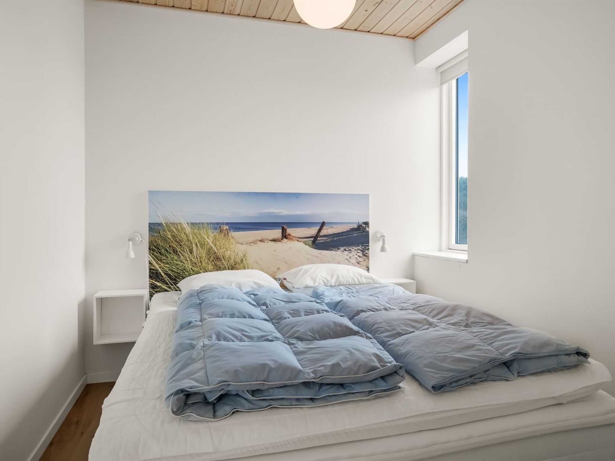 Un dormitorio con cama, mesita de noche, ventana y una pintura con vista a la playa.