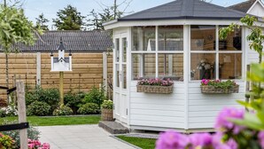 Eine weiße Laube mit Fenstern und Blumenkörben, umgeben von einem Garten und einem Holzzaun.