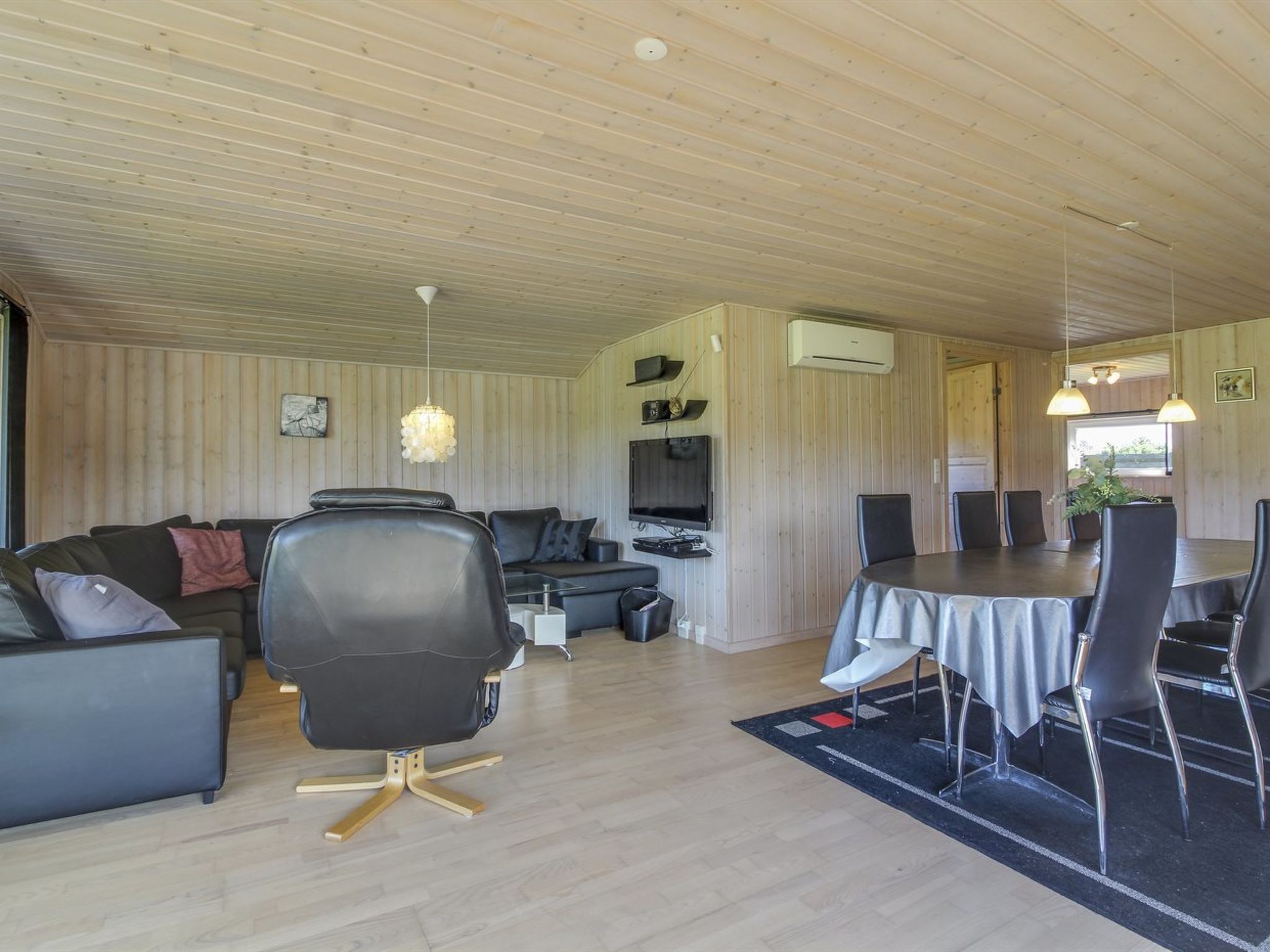 Ein modernes Wohnzimmer mit einer Couch, einem schwarzen Sessel, einem Esstisch mit Stühlen, einem Fernseher und einer Klimaanlage.