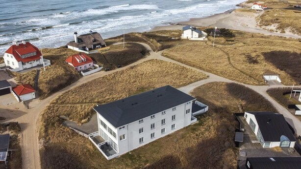 An aerial view of a coastal area with several buildings, including a large white building with a flat roof and multiple windows, surrounded by grassy fields and a beach.