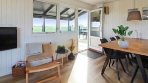 A room with a wooden dining table, chairs, a television, a wicker chair, a potted plant, and a view of a green field through the window. - "Lok" - 450m from the sea by Interhome (Løkken)