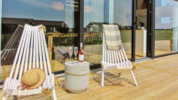 A deck with two white lounge chairs, a round stone table, and a bottle of wine.