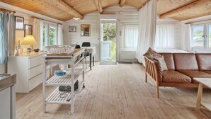 L’intérieur d’un chalet accueillant, avec un plafond en bois, un coin repas comprenant une table et des chaises, un canapé et une vue sur l’extérieur par une porte.