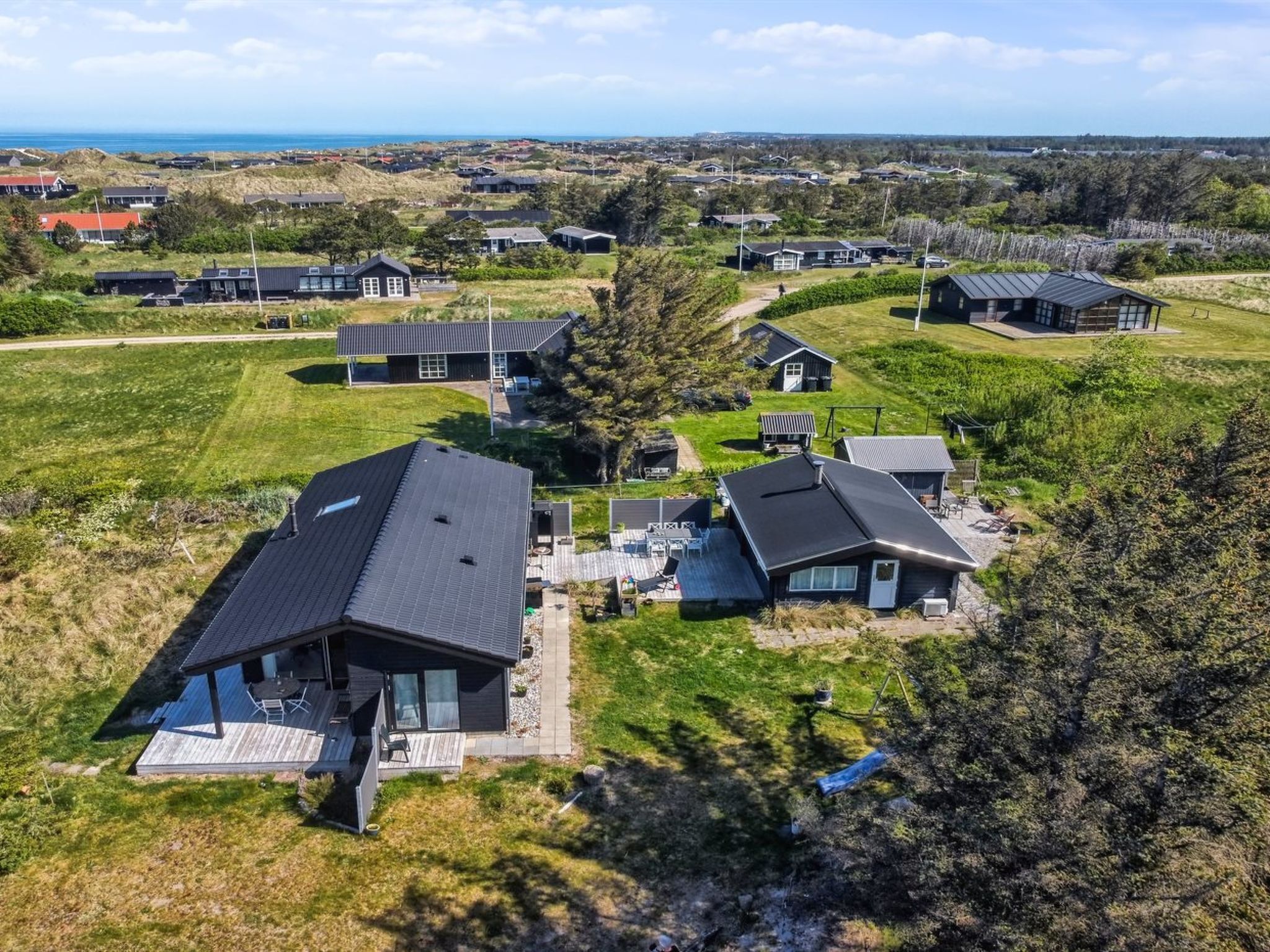 Vista aérea de uma área residencial com casas modernas, paisagem gramada e uma vista distante do mar.