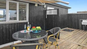Une terrasse avec une table ronde et deux chaises, une petite table avec des boissons et des fleurs, et une pergola au-dessus.