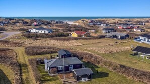 Vue aérienne d’un quartier résidentiel situé sur la côte, avec des maisons, un champ d’herbe et une plage en arrière-plan.