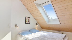 Un dormitorio con techo inclinado, tragaluz, cama con ropa de cama blanca, almohada azul y un cuadro en la pared.