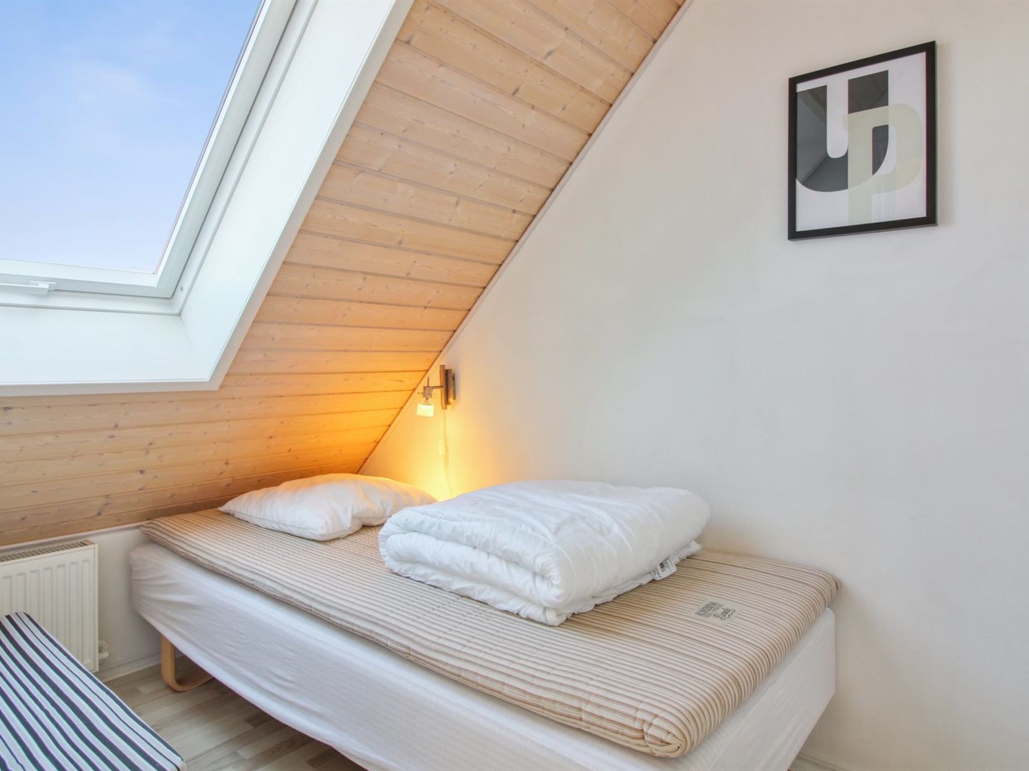 Un dormitorio con techo inclinado, tragaluces, cama con ropa de cama blanca, lámpara fijada a la pared y un cuadro enmarcado.