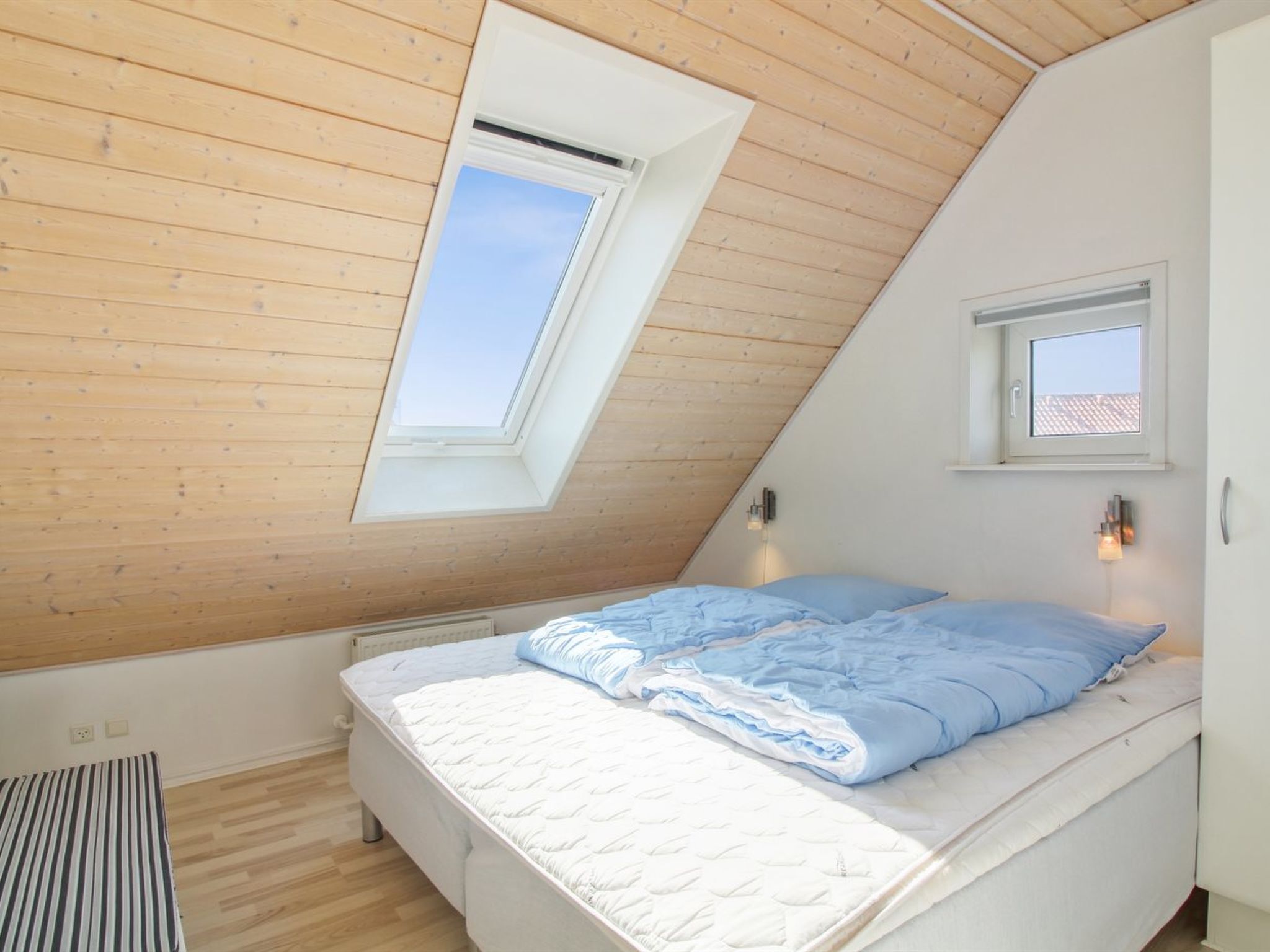 Une chambre avec un plafond en pente, un puits de lumière, un lit recouvert d’une couverture bleue et une petite fenêtre.