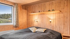 Une pièce aux murs lambrissés, comprenant un lit, deux lampes fixées au mur, une étagère ornée d’objets décoratifs et une fenêtre donnant sur un paysage verdoyant.