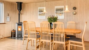 Une pièce aux murs lambrés, avec une table au centre, six chaises, un poêle à bois et deux lampes fixées au mur.
