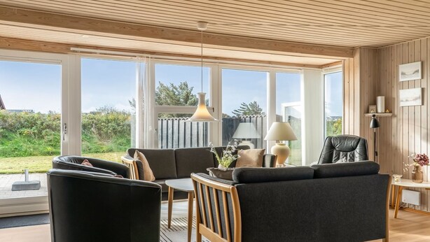 Un salon moderne avec un canapé, des fauteuils et une table à manger. De grandes fenêtres offrent une vue sur l’extérieur.