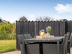 Un área de comedor al aire libre con una mesa negra y sillas de mimbre, una jarra de bebida anaranjada y un pequeño florero con flores.