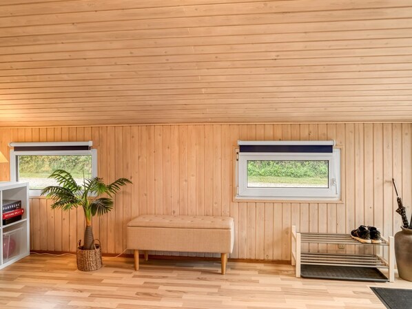Una habitación con paneles de madera, un banco, una planta, una estantería y dos ventanas.