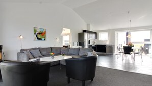 Una sala de estar moderna con un sofá seccional gris, una mesa de centro y sillones negros. Se ve una cocina al fondo, con un comedor visible a través de una puerta abierta.