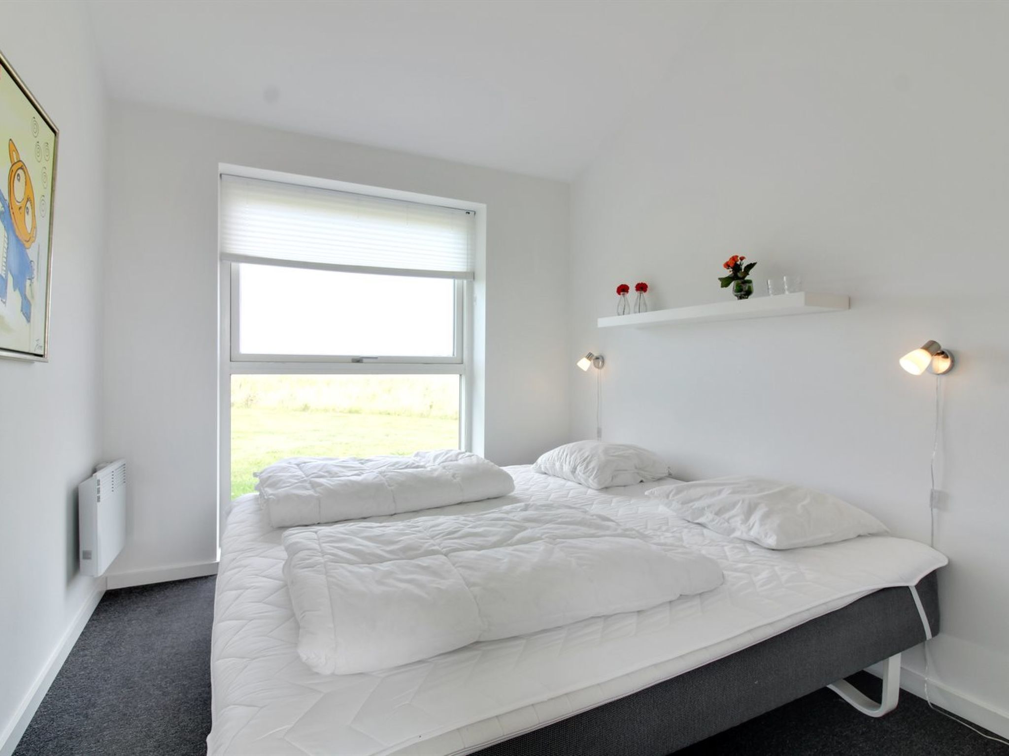 Un dormitorio con cama, ventana, radiador y estante en la pared con flores.