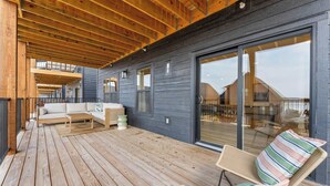 Una terraza de madera con sofá y sillas, puertas correderas de vidrio amplias, y vista al interior.
