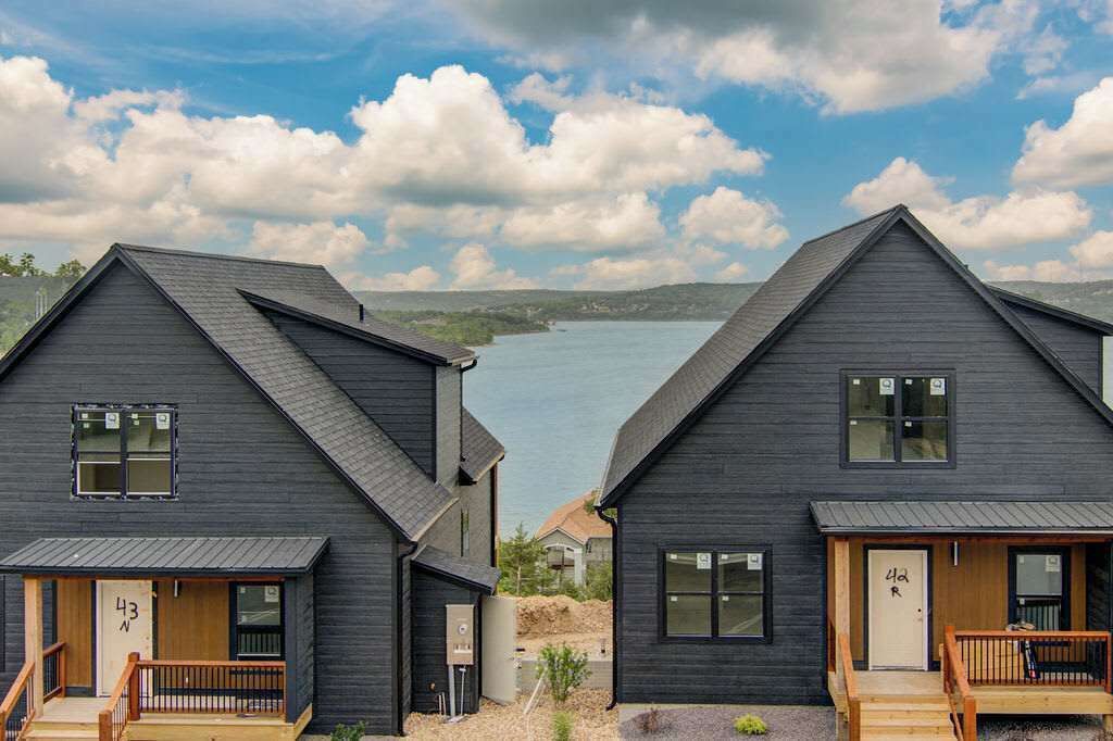 Two dark-colored houses with pitched roofs, wooden front porches, and a view of a lake.