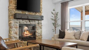 Une cheminée en pierre avec un téléviseur au-dessus, un canapé avec des coussins et une vue sur l’extérieur par une fenêtre.