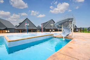 A swimming pool with a water slide, surrounded by buildings with grey roofs and a clear sky.
