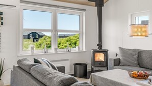 Ein modernes Wohnzimmer mit großem Fenster, einem schwarzen Kaminofen, grauen Sofas und einem Tisch mit Obstkorb.