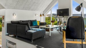 Un salon moderne avec un canapé gris, une table basse et un fauteuil noir. On y trouve un téléviseur posé sur un meuble et une fenêtre donnant sur l’extérieur.
