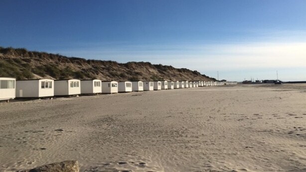 Une rangée de cabanes de plage blanches, dotées de fenêtres, est disposée le long d’une plage de sable, avec une dune en arrière-plan.