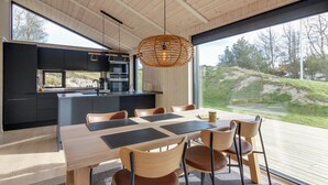 A modern kitchen with black cabinets and a large island, a dining area with a wooden table and chairs, and a large window offering a view of the outdoors.