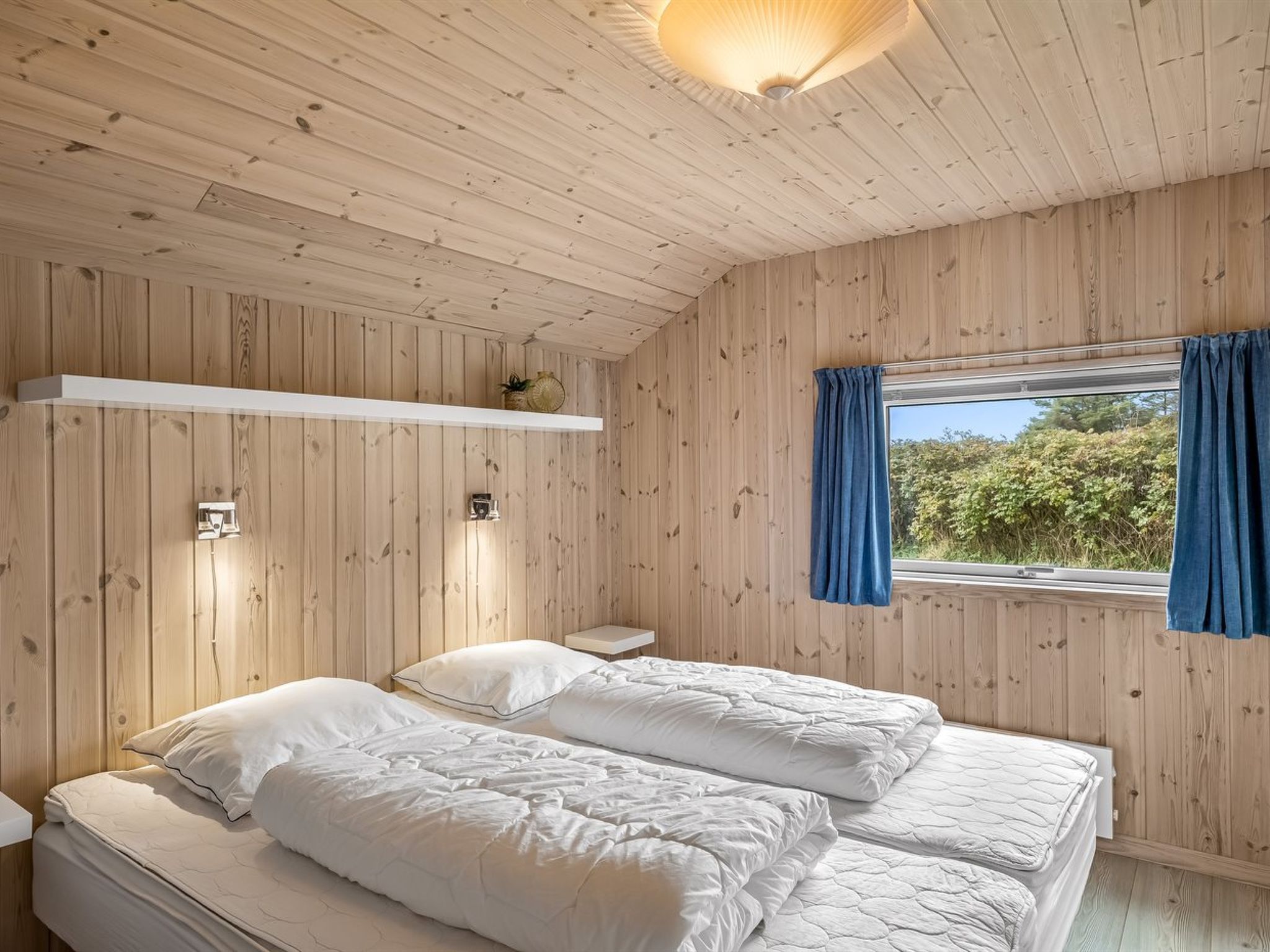 Ein Zimmer mit Holzwänden und -decke, zwei Betten mit weißer Bettwäsche, eine kleine Ablagefläche mit Dekoration und ein Fenster mit blauen Vorhängen.
