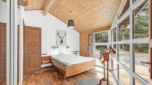 A modern bedroom with a wooden ceiling, a large bed, a desk, and a view of the outdoors through large windows.